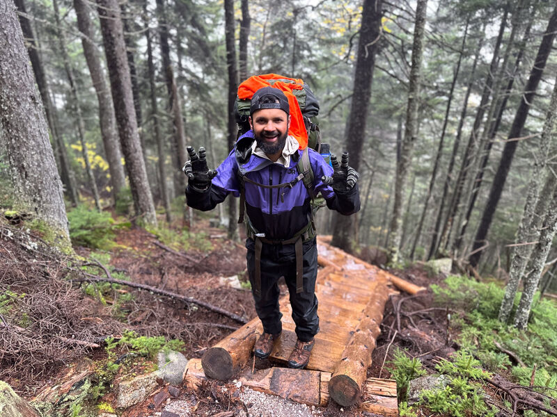 The image shows a man hiking in a forest. He is wearing a backpack, a jacket, and gloves. He is standing on a wooden bridge and giving a peace sign. The forest is dense with tall trees and the ground is covered in leaves and moss.